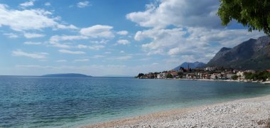 Im Vordergrund befinden sich ein Kies Strand und das blaue Meer. Am Horizont ist ein Dorf zu sehen.