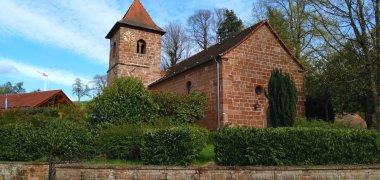 Kleine rote Sandsteinkirche, die durch eine Hecke eingesäumt wird.