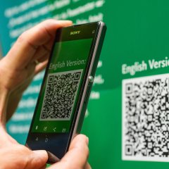 Grünes Informationsschild von dem ein QR-Code mit dem Handy abgescannt wird.