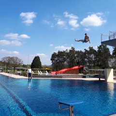 Ein Junge der von dem drei Meterbrett in den Pool springt. Neben dem Swimmingpool steht ein Mann und beobachtet den Jungen.