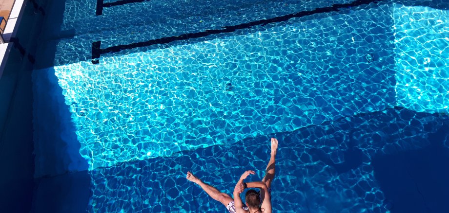 Ein junger Mann der breitbeinig in ein Schwimmbecken springt.