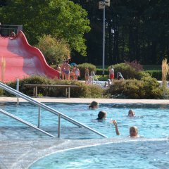 Fünf Menschen schwimmen im Schwimmbecken. Im Hintergrund ist eine breite rote Rutsche, auf der ein Junge steht und zwei sitzen. Weitere Menschen stehen an der Rutsche an.